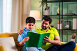 © StockImageFactory - Indian asian son reading a book with father on sofa while preparing for school lessons