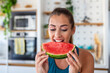 © Graphicroyalty - Young woman eating a slice of fresh watermelon in a modern kitchen. Happy female enjoying a healthy snack, biting into juicy red fruit, focusing on summer nutrition and a balanced organic diet.