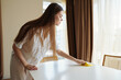 © SHOTPRIME STUDIO - woman cleaning table cloth interior chair curtains sunlight in a bright home scene, wiping dining surface with yellow cloth during morning routine, tidy apartment interior and cozy domestic care