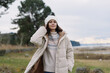 © SHOTPRIME STUDIO - Young woman in a warm beige winter coat stands outdoors by a lakeside, wearing a knit sweater and beanie in a breezy rural landscape today.