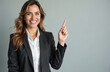 © miss irine - Smiling business woman in black suit points index finger up. Female leader suggests idea, indicates upward trend. Confident executive in office wear shows direction.