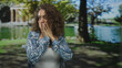 © Krakenimages.com - Woman with hands on face in park by a lake, holding palms to cheeks and covering mouth while standing outdoors; quiet concern.