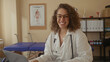 © Krakenimages.com - Woman doctor in white coat with stethoscope and glasses smiles while typing on laptop at clinic desk with therapy table behind; compassion care trust.