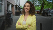 © Krakenimages.com - Woman smiling with arms crossed on a street near building and tree, wearing yellow shirt and tank top; confidence empowerment.
