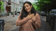 © Krakenimages.com - Young hispanic woman clasping hands to chest on a residential street with parked cars and bicycles; gratitude warmth.