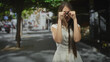 © Krakenimages.com - Young woman with glasses and long hair in white dress raises hands and rubs eyes on street; confusion introspection.