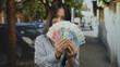 © Krakenimages.com - Young chinese woman outdoors on a street smiling while pointing at a fan of chinese yuan banknotes, showcasing money happiness and financial success in an urban setting.