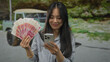 © Krakenimages.com - Young woman in park holding yuan banknotes and a smartphone outdoors, smiling in a green setting suggesting financial success.