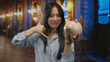© Krakenimages.com - Young chinese woman holding piggy bank and making a call me gesture while standing in a museum indoors beautifully illuminated with elegant decor.