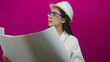 © Krakenimages.com - Young chinese woman architect studies blueprints wearing hardhat against isolated pink background, focusing intently on construction plans with a vibrant wall enhancing the scene.