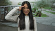 © Krakenimages.com - Young chinese woman smiling and saluting in a green park setting with trees and paved path, wearing casual white top outdoors with blurred background.