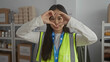 © Krakenimages.com - Young woman in a reflective vest makes a heart gesture at a volunteer center, surrounded by charity supplies, emphasizing community and kindness.