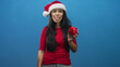 © Krakenimages.com - Woman in red top wearing santa hat holds a red piggy bank at chest level and sticks tongue out toward camera in a blue studio; holiday saving playful.