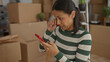 © Krakenimages.com - Woman using smartphone in new apartment surrounded by moving boxes showing a young hispanic woman indoors engaged with her phone during unpacking in her new home.