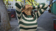 © Krakenimages.com - Hispanic woman in a striped shirt expressing frustration on a busy outdoor street with blurred urban background and sunlight filtering through trees.