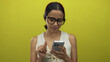 © Krakenimages.com - Young hispanic woman using smartphone against a vibrant yellow background, wearing glasses and a sleeveless top, displaying a cheerful expression as she interacts with her device.
