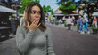 © Krakenimages.com - Young hispanic woman covering mouth with hand while looking sideways on busy street market; surprise secrecy.