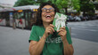 © Krakenimages.com - Middle-aged hispanic woman volunteer joyfully holding a gift box outdoors, with a city bus in the background, wearing a green volunteer shirt and smiling broadly in the street.