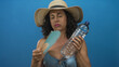 © Krakenimages.com - Hispanic middle-aged woman in bikini cools with hand fan, holding water bottle against vibrant blue background, conveying summer heat and refreshment.