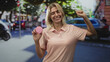 © Krakenimages.com - Woman holding a pink macaron with raised arm on a busy city street, smiling broadly; playful happiness.