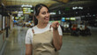 © Krakenimages.com - Woman wearing beige apron beckoning with hand in busy airport terminal near seating area and departure monitors; friendly welcome.