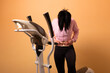 © alesmunt - Young woman in sportswear clutching her stomach beside an elliptical machine against an orange background, illustrating discomfort or fitness challenges.