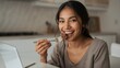 © LimeSky - Joyful young woman savoring cake with a fork, seated at a laptop with a notebook and pen, delighting in each bite while gazing at the camera in a kitchen setting