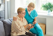 © Studio Romantic - Kind caregiver nurse, senior woman sitting on sofa in home, older patient pointing at smartphone, supportive and attentive young doctor, digital assistance learning during home care or personal helper