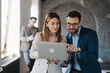 © Novak - Two focused business colleagues analyzing data on a laptop screen while a coworker works in the background of a modern office.
