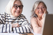© luciano - Two smiling senior women with white hair and glasses sitting at a wooden table at home, looking at a laptop together. They appear relaxed and happy while browsing online, with papers and a coffee mug