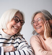 © luciano - Two smiling senior women with white hair and glasses sitting at a wooden table at home, looking at a laptop together. They appear relaxed and happy while browsing online, with papers and a coffee mug