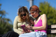 © Nikola Spasenoski - Two women sitting on a bench outside, using a phone and sharing a moment in bright sunlight on a clear day in a public park