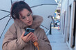 © tetxu - Woman standing in street holding umbrella looking at smartphone