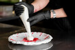 © pavel siamionov - A pastry chef wearing black gloves uses a white piping bag to decorate a dessert plate with cream over a vibrant red sauce splash in a professional kitchen