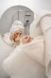 © Andrii Lysenko - Middle aged woman in bathrobe and towel applying moisturizing eye patches in bathroom mirror for skin care routine.