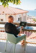 © Soloviova Liudmyla - Freelancer bald fency man enjoying remote work on sunny terrace surrounded by greenery with sea view. Digital nomad using laptop embracing freedom flexible lifestyle in warm tropic island climate