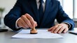 © Fairy Art - A close-up of a businessman in a dark blue suit stamping official documents with a wooden rubber stamp on his desk.