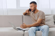 © SHOTPRIME STUDIO - Productive man talking on the phone while working on a laptop at home. This modern lifestyle representation highlights remote work and effective communication.