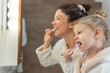 © lordn - Smiling mother and young daughter in bathrobes brush their teeth together in a bathroom mirror, showing daily hygiene routine, positive parenting, learning by example, and warm family bonding at home.