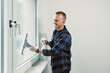 © DSMT - Man cleans window in bright room during daytime with a smile while using a squeegee and cloth to remove dirt and spots from glass