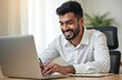 © Viktor - Smiling Indian man works on laptop in office. Young businessman types on computer, takes notes, learns online. Focused pro with beard at desk, busy with work tasks.