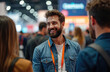 © Maryna - Man smiles at trade fair in conversation with colleagues. Professionals network at busy convention hall, discuss ideas. Businesspeople gather at expo event.