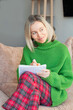 © Tatyana Gladskih - Smiling Woman sitting on Sofa in living room and making Wish list for next year, Female in green sweater writes down shopping list in notebook while sitting at home