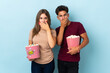© luismolinero - Couple eating popcorn while watching a movie isolated on purple background covering mouth with hands for saying something inappropriate