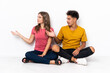 © luismolinero - Young couple sitting on the floor isolated on white background extending hands to the side for inviting to come