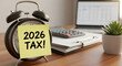 © Utpol - A vintage black alarm clock with a yellow sticky note displaying '2026 TAX!' on a wooden desk, alongside a blurred laptop, calculator, and small potted plant