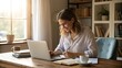 © shameer - A young woman working on her laptop and taking notes in a cozy home office with natural light