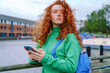 © Iryna - Young woman with curly red hair uses smartphone by the canal in a city on a cloudy day