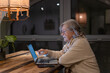 © luciano - Senior woman using a laptop at home in the evening, sitting at a wooden table under warm light, representing digital literacy, online communication, and independent lifestyle for older adults