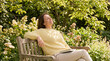 © OlgaNeuroArt - Middle aged woman relaxing on a wooden bench in a sunny garden. Happy female sitting outdoors with a book on her lap. Wellness and mindfulness concept